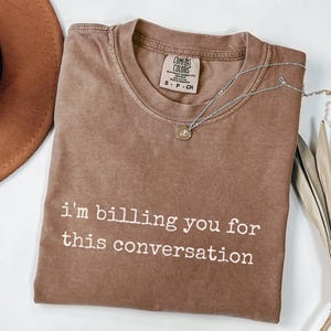 May include: A brown Comfort Colors t-shirt with the text "I'm billing you for this conversation" in white. A silver necklace with a small pendant is on the shirt. A brown hat and dried plant are in the background.