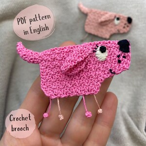 May include: A handmade pink crochet dog brooch with black and white embroidered details. The dog has dangling legs and a smiling face. The image also includes a smaller, similar brooch and text that says "PDF pattern in English" and "Crochet brooch".