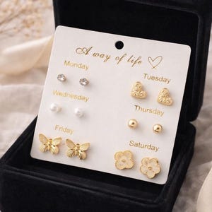 May include: A black velvet jewelry box holds a set of gold-colored earrings. The earrings are arranged on a white card with the words "A way of life." The earrings include studs, pearls, hearts, butterflies, and flowers.