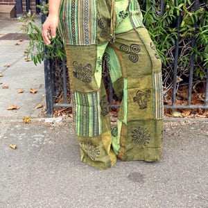 May include: Olive green patchwork trousers featuring an elastic waistband with a drawstring. The trousers are constructed from various fabric panels in shades of green, decorated with black printed motifs including mushrooms, spirals, and flowers.