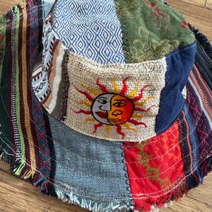 May include: A patchwork bucket hat featuring a variety of colourful fabric panels including blue diamond patterns, green floral prints, and red geometric designs. The front panel displays an embroidered sun and moon face with a frayed brim edge.