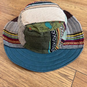 May include: A patchwork bucket hat featuring a variety of fabric textures and patterns, including a cream-coloured diamond-patterned top, olive green panels, a dark teal brim, and horizontal bands of red, yellow, blue, and black woven designs.