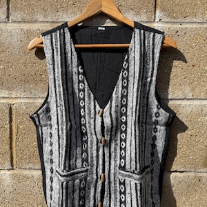 May include: A grey and black striped waistcoat with a V-neck and wooden buttons. The waistcoat has two front pockets and is hanging on a wooden hanger. The waistcoat is made of a woven fabric.