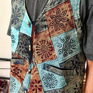 May include: A sleeveless waistcoat featuring a patchwork design of blue, brown, and black fabric squares. Each square is decorated with a dark, stamped floral or sun-like pattern. The waistcoat includes four light-coloured wooden toggle buttons for closure.