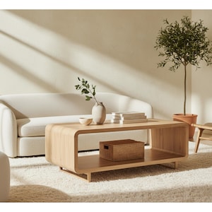 May include: A modern living room scene featuring a light wood coffee table with rounded edges, a white sofa, and a woven storage box. A small tree in a terracotta pot and a woven chair add to the room's decor.