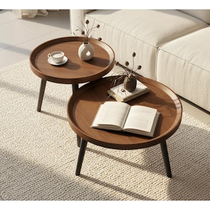 May include: Two round, wooden side tables with dark legs. One table has a cup and saucer, and a small vase. The other table has a book and a small vase. The tables are on a cream-colored rug.