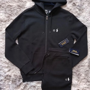 Puede incluir: Conjunto de chándal negro con capucha, que incluye una chaqueta con cremallera y un pantalón a juego. La chaqueta tiene un pequeño logotipo en el pecho y bolsillos delanteros. El pantalón tiene cintura con cordón y un logotipo a juego. Ambos artículos tienen etiquetas.