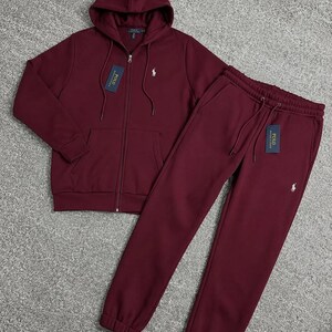 May include: A burgundy tracksuit set, featuring a zip-up hoodie with a drawstring hood and matching sweatpants. Both pieces have a small white logo on the chest and leg. The set includes tags.