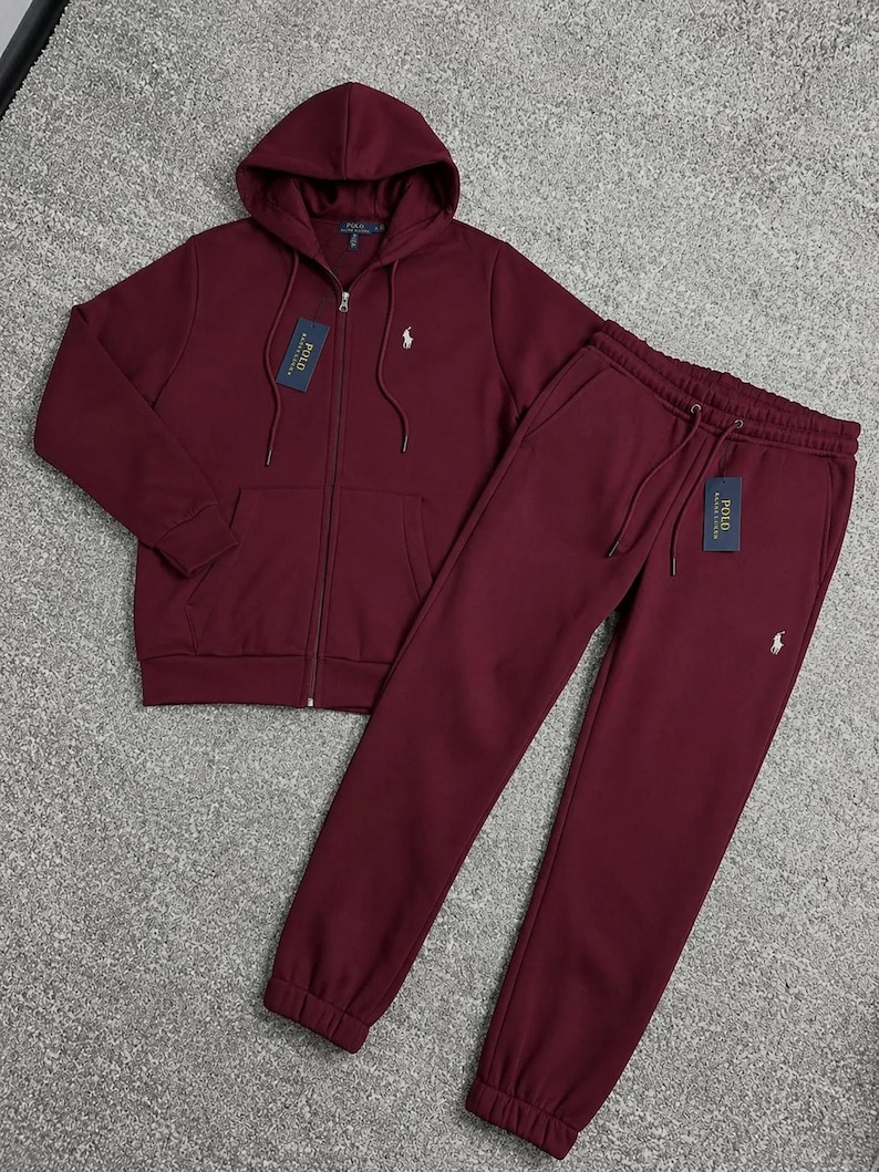 May include: A burgundy tracksuit set, featuring a zip-up hoodie with a drawstring hood and matching sweatpants. Both pieces have a small white logo on the chest and leg. The set includes tags.