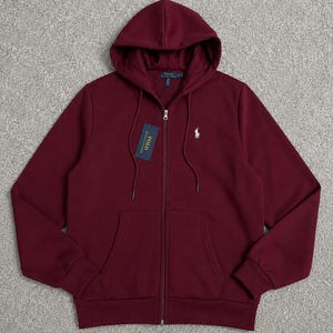 May include: A burgundy zip-up hoodie with a drawstring hood and front pockets. The hoodie features a white logo on the chest and a tag with text. A casual, comfortable garment.