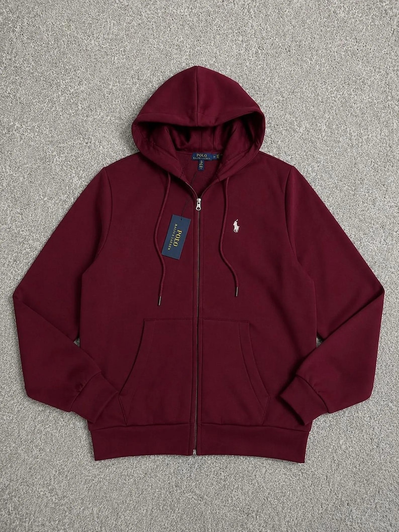 May include: A burgundy zip-up hoodie with a drawstring hood and front pockets. The hoodie features a white logo on the chest and a tag with text. A casual, comfortable garment.