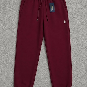 May include: Burgundy-colored sweatpants with a drawstring waist and elastic cuffs. A small white logo is on the left leg, and a tag is attached to the waistband. The pants have two side pockets and are made of a soft material.