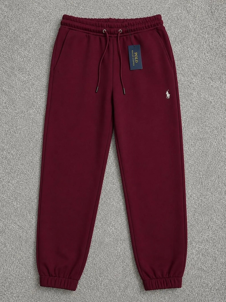 May include: Burgundy-colored sweatpants with a drawstring waist and elastic cuffs. A small white logo is on the left leg, and a tag is attached to the waistband. The pants have two side pockets and are made of a soft material.
