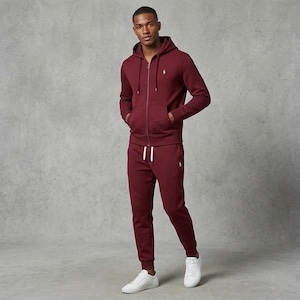 May include: A burgundy zip-up hoodie and matching sweatpants set. The hoodie has a drawstring hood and a small logo on the chest. The sweatpants have a drawstring waist and are cuffed at the ankles. The outfit is paired with white sneakers.