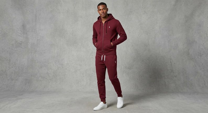 May include: A burgundy zip-up hoodie and matching sweatpants set. The hoodie has a drawstring hood and a small logo on the chest. The sweatpants have a drawstring waist and are cuffed at the ankles. The outfit is paired with white sneakers.