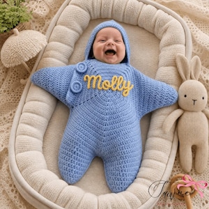 May include: A light blue crocheted star-shaped baby bunting with a hood, featuring two matching blue buttons on the upper chest and the name Molly embroidered in yellow cursive script across the center of the chest area.