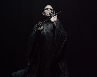 May include: A detailed figurine of a pale, bald character with a snake-like nose, wearing a long, flowing black robe. The figure holds a thin, dark wand in one hand, positioned near the face, standing atop a base shaped like an open book.