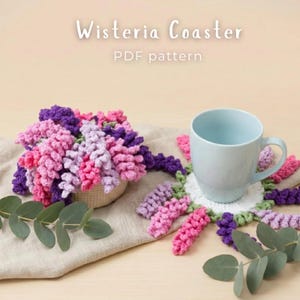 Puede incluir: Un patrón PDF de posavasos Wisteria de ganchillo con una taza azul claro sobre un posavasos en forma de flor blanca, verde y morada. Al lado hay un arreglo floral de ganchillo a juego en un recipiente pequeño con hojas verdes.