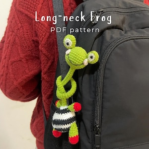 May include: A crocheted long-neck frog toy with a green head and body, white and black striped belly, and red accents on the hands and feet. The frog is wrapped around a black backpack strap. Text reads: Long-neck Frog PDF pattern.