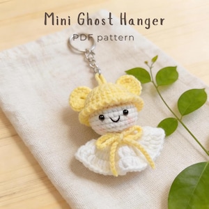 May include: A crochet keychain featuring a white ghost figure wearing a yellow hat and a yellow bow. The ghost has black bead eyes and a stitched smile. The text Mini Ghost Hanger PDF pattern is visible above the keychain on a light fabric.