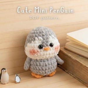 May include: A crocheted penguin plush with gray and white yarn, black plastic eyes, an orange beak, and orange feet, featuring pink blush on its cheeks. The text "Cute Mini PenGuin PDF pattern" is displayed above the plush toy.