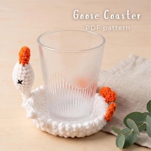Peut inclure: Un dessous de verre en crochet blanc en forme d'oie avec un bec et des pattes orange, sur lequel est posé un verre transparent strié. Le texte Goose Coaster PDF pattern est visible en haut, indiquant un document numérique pour ce projet.