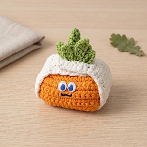 May include: A handmade crochet plush toy shaped like a radish. The radish is orange with a white crocheted wrap and green leafy top. It has googly eyes and a black embroidered mouth. The toy is on a light wood surface.