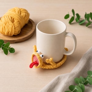 Könnte beinhalten: Eine cremefarbene Tasse steht auf einem gelben, gehäkelten Untersetzer in Form eines Monsters mit roter Zunge. Ein Knäuel gelber Wolle liegt auf einem Holztablett. Grüne Blätter sind um die Tasse verstreut.