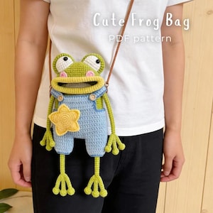 Könnte beinhalten: Eine gehäkelte Froschtasche mit braunem Riemen, die einen grünen Frosch in einer hellblauen Latzhose mit gelbem Sternaufnäher zeigt. Die Tasche hat weiße Augen mit schwarzen Pupillen und rosa Wangen. Text "Cute Frog Bag PDF pattern" ist sichtbar.