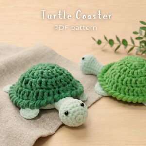 Pode incluir: Dois porta-copos de crochê em formato de tartaruga com cascos verde-escuros e cabeças e membros verde-claros. O texto "Turtle Coaster PDF pattern" é visível no topo. As tartarugas possuem pequenos olhos de contas pretas e pontos texturizados.