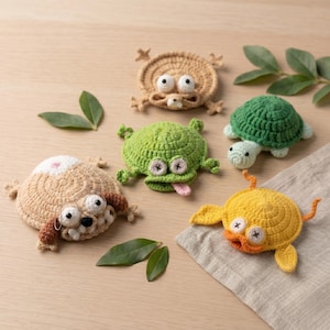 Peut inclure: Ensemble de dessous de verre animaux crochetés à la main. L'ensemble comprend un chien, une grenouille, une tortue, un canard et un animal rond avec de grands yeux. Les dessous de verre sont dans les tons de marron, vert, jaune et blanc, avec des accents noirs et blancs. La grenouille a une langue rose.