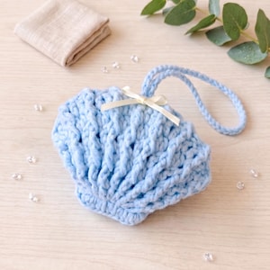 Puede incluir: Un jabonera en forma de concha de ganchillo azul claro con un asa y un pequeño lazo color crema. En el fondo, un paño de lavado beige doblado. La jabonera está diseñada para contener una pastilla de jabón.