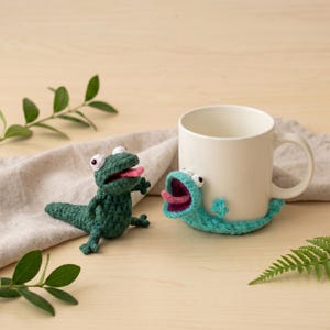 May include: A white ceramic mug with a crocheted teal monster attached, and a separate green crocheted monster. Both have open mouths with pink tongues and white eyes. The items are on a wooden surface.