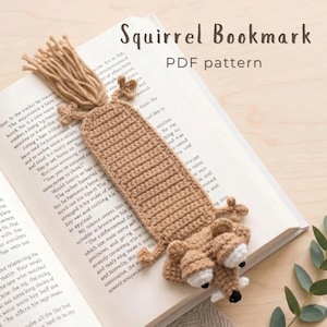 May include: A light brown crocheted squirrel bookmark with a long tail, small limbs, and a head featuring white and black eyes. The bookmark is placed on an open book. The text "Squirrel Bookmark PDF pattern" is printed at the top of the image.