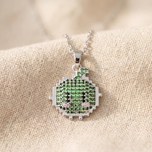 May include: A silver-toned pendant necklace featuring a pixelated, square-shaped character design. The pendant is encrusted with small green stones, two black stones for eyes, and two small pink stones for cheeks, suspended from a delicate silver chain.