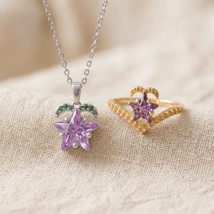 Peut inclure: Un collier argenté avec un pendentif en forme d'étoile violette et un accent incurvé serti de petites pierres vertes, accompagné d'une bague chevron dorée avec une étoile violette centrale et des détails métalliques texturés.