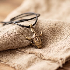 Può includere: Un ciondolo in metallo color bronzo a forma di volto umano con corna ricurve, sospeso a un sottile cordino nero. Il ciondolo presenta tratti del viso dettagliati e un motivo decorativo sulla fronte, con una finitura metallica liscia.