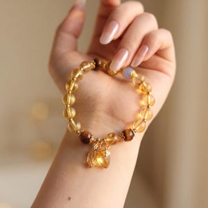 Pode incluir: Uma pulseira de contas com esferas de citrino amarelo translúcido, contas de olho de tigre castanho escuro e uma conta de cor lavanda pálida. A pulseira inclui espaçadores de metal dourado e dois pingentes dourados em forma de folha.