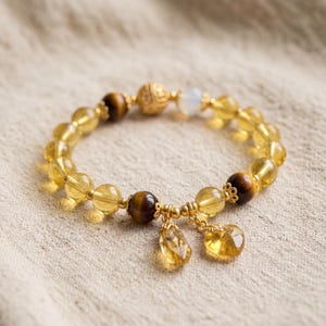 Puede incluir: Una pulsera de cuentas con esferas de citrino amarillo translúcido, cuentas de ojo de tigre marrón y una cuenta de piedra lunar blanca. El diseño incluye espaciadores de metal dorado, una cuenta dorada texturizada y dos dijes de citrino.
