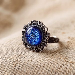 Puede incluir: Un anillo de tono plateado con un cabujón circular de color azul intenso en el centro que presenta un patrón radiante similar a una estrella. El engaste es ornamentado con intrincados detalles metálicos en espiral y la banda tiene textura.