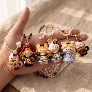 Puede incluir: Una colección de seis collares con figuras en miniatura de conejos y gatos vestidos con trajes elaborados, incluyendo una reina, un invitado a una fiesta de té y un personaje con vestido azul, suspendidos en delicadas cadenas de cuentas doradas.