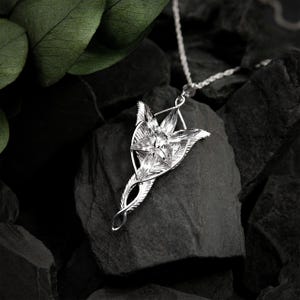 May include: A silver pendant necklace with a star-shaped design and a central crystal. The pendant hangs from a fine silver chain. The necklace is displayed against a dark, textured backdrop, with a leaf in the background.