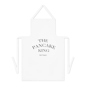 Puede incluir: Un delantal blanco con el texto "THE PANCAKE KING" en negro, con una fuente elegante. Debajo, se lee "Get Flippin". El delantal tiene un lazo para el cuello y correas laterales.
