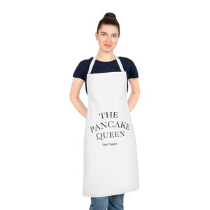 Puede incluir: Un delantal blanco con el texto "THE PANCAKE QUEEN" en letras negras. Debajo del texto principal está "Get Flippin". El delantal tiene una correa para el cuello y lazos en la cintura.