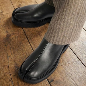 May include: Black leather split-toe shoes with a thick, textured sole. The shoes have a unique design with a separation for the big toe. The shoes are worn with brown patterned trousers, creating a modern, stylish look.