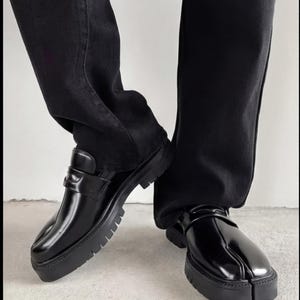 May include: Black leather loafers with a split-toe design and thick, lugged soles. The shoes have a glossy finish and a strap detail across the top. The image shows the shoes paired with black denim jeans.