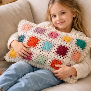 May include: A rectangular, handmade crochet pillow with a colorful patchwork design. The pillow features squares of various colors, including orange, pink, blue, and yellow, set against a cream-colored background. The edges are finished with a ruffled trim.