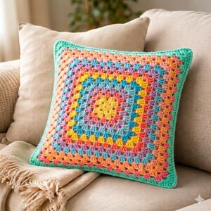 May include: A square, handmade crochet pillow with a vibrant, multicolored design. The pillow features a granny square pattern in shades of yellow, orange, pink, blue, and purple, framed by a green border. It rests on a beige sofa, suggesting home decor.