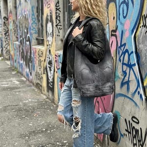 May include: A person wearing a black leather jacket, black shirt, and light blue ripped jeans, carrying a gray shoulder bag. The background features a graffiti-covered wall with colorful artwork and text.