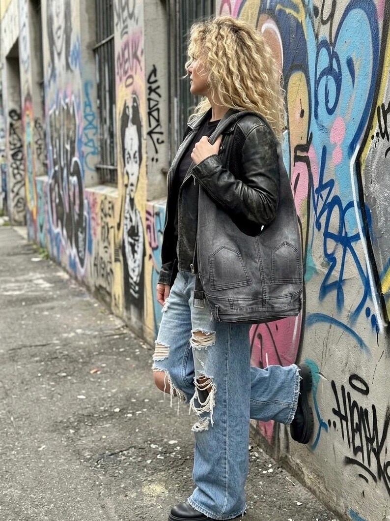 May include: A person wearing a black leather jacket, black shirt, and light blue ripped jeans, carrying a gray shoulder bag. The background features a graffiti-covered wall with colorful artwork and text.